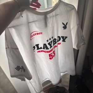 Vit Playboy Racing Club t-shirt - Vit t-shirt från Playboy med tryck i svart och rött framtill, där det står 'Playboy Racing Club 53'. Har korta ärmar med schackrutiga flaggor på ena ärmen och klassisk Playboy-kanin på bröstet. Skön och avslappnad passform, perfekt för en cool streetstyle.