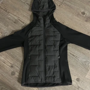 Svart quiltad pufferjacka med huva ( vadderad) - Svart pufferjacka med quiltade paneler fram och bak, hel dragkedja och huva. Jackan har långa ärmar och två sidofickor med dragkedja. Perfekt för kyliga dagar och har en sportig, clean look, funkar även till ridning.
