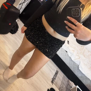 Svart paljettkjol i minilängd - Supersnygg svart minikjol täckt med glittrande paljetter. Kjolen har en tight passform och sitter högt i midjan, perfekt för att sticka ut. Den är kort och har en klassisk rak modell som ger en trendig look.