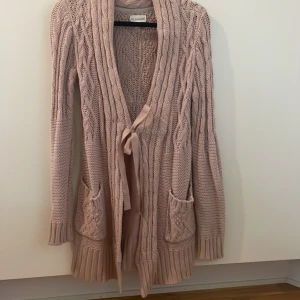 Ljusrosa stickad kofta från 365 Sunshine - Mysig gammelrosa stickad kofta från 365 Sunshine med kabelstickat mönster, två fickor fram och ribbade muddar. Koftan har lång ärm och knyts med ett dekorativt band.  Storlek XS men passar även S. 