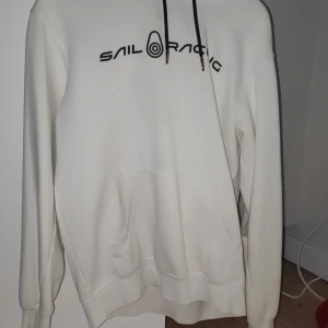 Vit hoodie från Sail Racing - Vit hoodie från Sail Racing med svart logga framtill och tryck på huvan. Klassisk känguruficka, svarta snören och mjuk känsla. Perfekt för dig som gillar stilren och sportig look.