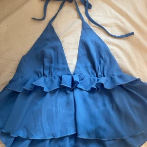 Blå volang topp med knytband - Säljer en blå ärmlös topp med djup v-ringning och volangdetaljer. Toppen har knytband i nacken och dubbla lager tyg som ger en flowy känsla. Perfekt för sommarens alla tillfällen och ger en trendig look.