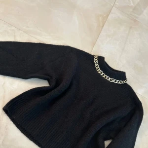 Svart stickad tröja med guldkedja H&M - Svart stickad tröja från H&M med lång ärm och ribbade muddar. Tröjan har en hög halsringning som är dekorerad med en chunky guldfärgad kedja för en extra edgy vibe. Perfekt för dig som vill sticka ut lite extra.