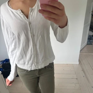 Vit kofta med knappar - Säljer en klassisk vit kofta med långa ärmar och knäppning framtill. Koftan är i ett tunt, mjukt material och har en enkel, tidlös design som passar perfekt till jeans eller kjol.