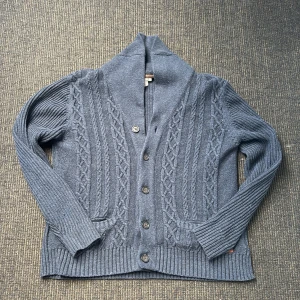 Mörkblå stickad kofta med mönster - Mysig mörkblå stickad kofta med snyggt kabelstickat mönster framtill, bred krage och knappar hela vägen. Två fickor framtill och ribbade muddar ger en chill och klassisk look. Perfekt för lager-på-lager och dessa kyliga dagar som kommer. Storlek L, fint skick. Tveka inte på att höra av er vid funderingar!