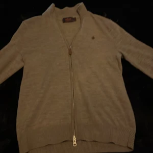 Beige kofta från Morris med dragkedja - Snygg beige kofta från Morris i mjuk ullblandning. Koftan har hel dragkedja framtill med läderdetalj och en broderad logga på bröstet. Perfekt för lager-på-lager och passar till det mesta. Klassisk och stilren design.