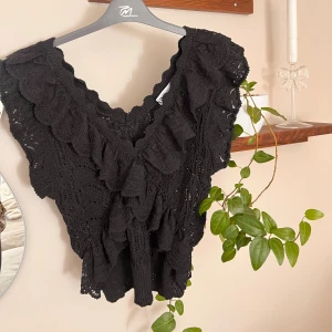 Svart spetsblus med volang från Zara - Supersöt svart blus från Zara med v-ringning och breda volanger längs halsringningen. Blusen är ärmlös och har ett detaljerat spetsmönster som ger en romantisk vibe. Perfekt att styla med jeans eller kjol för en trendig look.