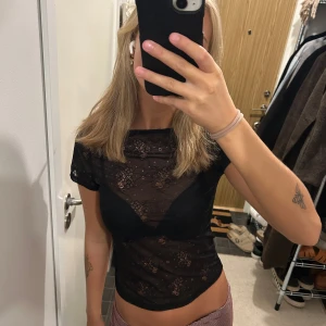 Svart spetsblus med öppen rygg - Säljer en svart, genomskinlig spetsblus med korta ärmar och snygg öppen rygg. Blusen har ett blommigt spetsmönster och är croppad i modellen. Perfekt att styla med ett linne eller snygg bh under.