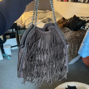 Brun axelväska med fransar och kedja - Stella Mccartney-liknande väska. Stor brun axelväska i mocka med coola fransar på framsidan och grov silverfärgad kedja som axelrem. Baksidan har en praktisk dragkedja. Perfekt för dig som gillar boho-vibe och vill ha plats för allt du behöver. Supbra skick🫶💕