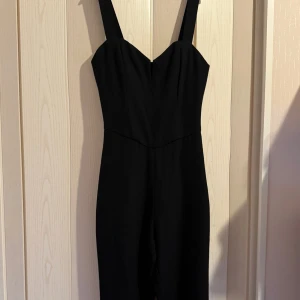 Svart långklänning med v-ringad urringning - Snygg svart långklänning med breda axelband och v-ringad urringning. Klänningen har en figurnära passform upptill och är tillverkad i ett slätt, vävt material med fodrad insida. Perfekt för dig som gillar stilrena och eleganta looks.