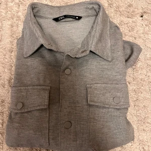Grå overshirt från Zara i bomull - Snygg grå overshirt från Zara med klassisk krage, två stora bröstfickor med knappar och långa ärmar. Overshirten är i mjuk bomull och har en clean look med tryckknappar framtill. Perfekt att slänga över en t-shirt för en chill vibe. Dock en jätte liten sprätt på trådar inte så mycket, ett litet hål. 
