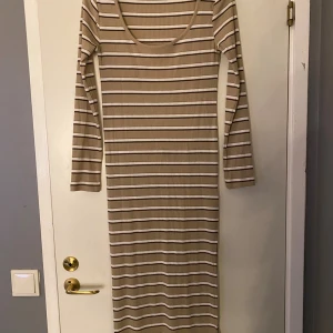 Beige och vit randig långklänning 157 - Superfin långklänning från 157 i ribbad beige och vit bomullsmix med svarta och bruna ränder. Klänningen har lång ärm, rundad ringning och sitter figurnära. Perfekt för dig som gillar en enkel men snygg stil.Frakt ingår inte 