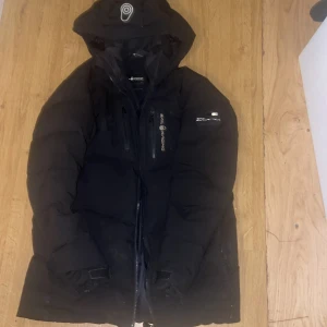 Svart dunjacka från Sail Racing - Svart dunjacka från Sail Racing med huva och flera dragkedjeförsedda fickor. Jackan har en stilren look med logga på bröstet och är tillverkad i vindtätt och andningsbart material. Perfekt för kalla dagar och har en skön, vadderad känsla.