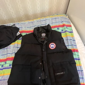 Svart dunväst från Canada Goose - Svart dunväst från Canada Goose med klassisk logga på bröstet och två stora fickor framtill. Västen har hög krage, quiltad design och stängs med både dragkedja och knappar. Perfekt för lager-på-lager och stilren look.