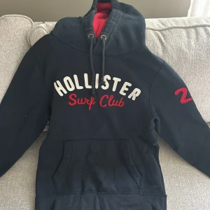 Svart hoodie Hollister Surf Club M - Svart hoodie från Hollister med broderad text 'Surf Club' i rött och vitt framtill samt röd siffra på ärmen. Stor huva med snörning och röd insida, samt stor magficka. Perfekt för dig som gillar en sportig och avslappnad stil.