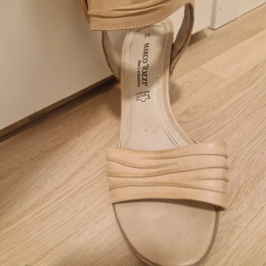 Beiga sandaler med klack från Marco Tozzi - Snygga beiga sandaler från Marco Tozzi i skinn med bred rem över tårna och dekorativa veck. Klack, öppen tå och dragkedja på sidan för enkel påtagning. Stort knapp-liknande dekordetalj vid ankeln. Perfekta för dig som gillar stilrena och bekväma skor.