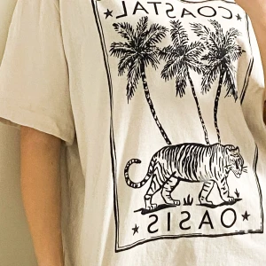 Beige oversized t-shirt H&M med tryck - Snygg beige t-shirt från H&M i oversized modell med svart tryck av palmer och tiger samt texten 'COASTAL OASIS' på framsidan. Tillverkad i 100% bomull och har korta ärmar. Perfekt för en chill och avslappnad stil.