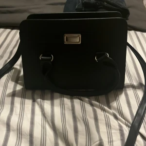 Svart fyrkantig axelväska i skinn - Stilren svart axelväska i fyrkantig modell med både handtag och axelrem. Väskan har silvriga metalldetaljer och flera fack, perfekt för att hålla ordning på dina grejer. Materialet är slätt och ger en clean look som passar till många outfits.