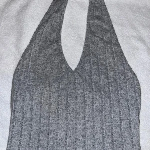 Grå ribbad halterneck linnetopp - Säljer en grå ribbad linnetopp med halterneck och djup v-ringning. Toppen är ärmlös och har en tight passform som framhäver axlarna. Perfekt för dig som gillar en enkel men trendig look. Helt ny