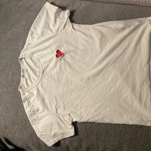 Vit t-shirt Comme des Garçons Play - Vit t-shirt från Comme des Garçons Play med det ikoniska röda hjärtat broderat på bröstet. Klassisk rund halsringning och korta ärmar. Tillverkad i mjuk bomull som känns skön mot huden. Perfekt för en clean och stilren look.