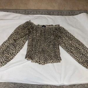 Blus - Trendig blus från Sisters Point i beige med svart leopardmönster. Modellen är offshoulder med långa, luftiga ärmar och smockad kropp som ger en snygg passform. Perfekt för dig som vill sticka ut med ett djurmönster och en cool siluett. Inga defekter