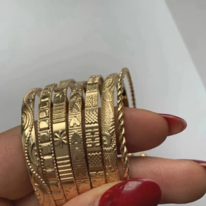 Guldiga armband med mönster 7 st - Säljer ett set med flera guldiga armband i olika bredder och mönster. Armbanden har detaljer som hjärtan, stjärnor, blommor och geometriska former samt ett tvinnat armband. Perfekt för dig som vill ha en unik och trendig accessoar till din outfit.