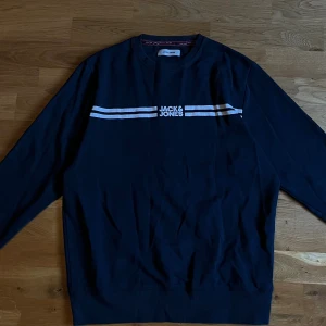 Svart sweatshirt från Jack & Jones - Svart sweatshirt från Jack & Jones med vita ränder och logga över bröstet. Klassisk rund halsringning och långa ärmar. Tillverkad i mjuk bomull, perfekt för en chill och stilren look.