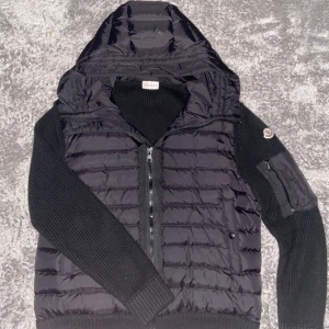 Svart cardigan med huva från Moncler - Sälja en svart dunväst från Moncler med stickade ärmar och huva. Västen har quiltad framsida, dragkedja, fickor och en snygg Moncler-logga på ärmen. Perfekt för lager-på-lager och riktigt stilren look.