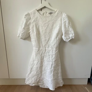 Vit kort klänning med broderade blommor - Superfin vit kort klänning från NA-KD med broderade blommor, puffärmar och öppen rygg med knappar. Klänningen är i bomull och har en söt, feminin look med markerad midja och lätt utställd kjol. Perfekt för sommarens alla tillfällen!