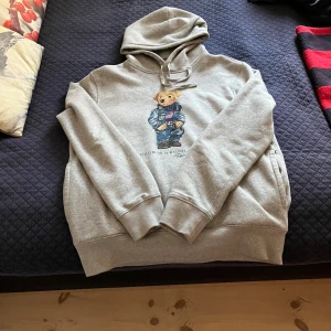 Grå hoodie Polo Ralph Lauren M - Säljer en grå hoodie från Polo Ralph Lauren i storlek M men passar även S, Tröjan har en den klassiska björnen på bröstet, snörning i huvan och klassisk känguruficka. Mjuk och skön bomullsblandning, perfekt för chill dagar. Använd 1-2 gånger inga skavanker eller defekter alls. Skriv för fler bilder eller om ni har frågor!