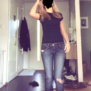 Mörkblå jeans med slitningar - Säljer ett par mörkblå jeans med snygga slitningar på låret och detaljerade bakfickor med kontrastsömmar. Jeansen har låg midja och klassisk femficksmodell. Perfekta för dig som gillar en avslappnad och trendig look.