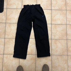 Svarta Nike täckbyxor med blå detalj - Svarta täckbyxor från Nike med blått innerfoder och blå Nike-logga på benet. Byxorna har elastisk midja med snörning för extra passform. Perfekta för kalla dagar och utomhusaktiviteter.