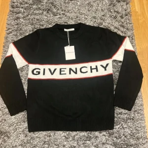 Svart stickad Givenchy tröja  - Svart stickad tröja från Givenchy med vit panel och röd kant över bröstet där loggan är broderad. Långa ärmar med vit detalj på axlarna och ribbade muddar. Perfekt för dig som vill ha en lyxig och stilren look.