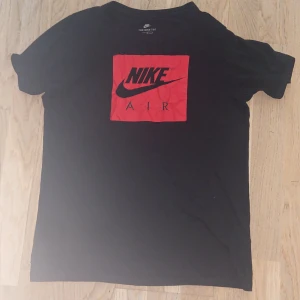 Svart Nike Air t-shirt med röd logga - Svart t-shirt från Nike med en stor röd fyrkantig logga och texten 'Nike Air' på bröstet. Klassisk rund hals och korta ärmar. Tillverkad i mjuk bomull som är skön att ha på sig. Perfekt för dig som gillar sportig och stilren streetwear.
