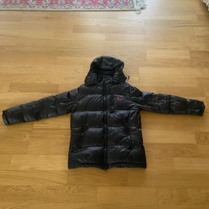 Svart dunjacka Ralph Lauren - Svart dunjacka från Ralph Lauren med huva och dragkedja framtill. Jackan har en quiltad design, två sidofickor med dragkedja och en bröstficka. Klassisk röd Polo-logga på bröstet. Perfekt för kalla dagar och riktigt snygg streetstil.