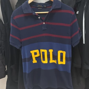 Randig piké från Polo Ralph Lauren - Snygg randig pikétröja från Polo Ralph Lauren i marinblått och vinrött med stora gula bokstäver 'POLO' framtill. Modellen är custom slim fit och har klassisk krage och korta ärmar. Perfekt för dig som gillar sportig och stilren look.