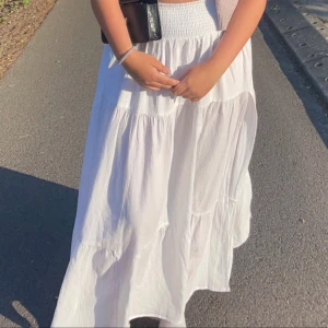 Vit långkjol med volanger - Superfin vit långkjol med volangdetaljer och smock i midjan. Kjolen är luftig och har en flowy känsla, perfekt för varma dagar. Passar snyggt till crop tops eller stickade tröjor.