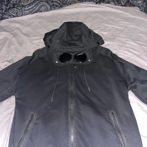 Svart gogglejacka från C.P. Company - Svart gogglejacka från C.P. Company med ikoniska glasögon i huvan. Tröjan är i nyskick och aldrig använd, säljer pga lite för stor på mig. Jackan har dragkedja framtill, två snedställda fickor med dragkedja och justerbar huva med snörning. Snygg och urban stil, perfekt för dig som gillar streetwear. Priset är inte hugget i sten, men tycker priset är bra satt jämfört me nypris och skicket den är i.
