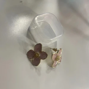 Blommiga örhängen i rosa och guld - Supersöta örhängen i form av blommor med fyra kronblad. De är rosa med gulddetaljer och har en glansig finish. Perfekta för att ge din look en söt och feminin touch. Kommer med praktisk liten ask för förvaring.