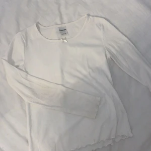 Vit långärmad topp med rosett Kappahl - Söt vit långärmad topp från Kappahl med liten rosett vid halsringningen. Tröjan har ribbad struktur och vågig kant nertill för en extra detalj. Perfekt för lager-på-lager och passar till allt. Liten fläck längst ner på vänstra armen (se bild)