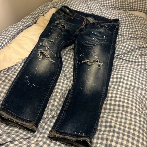 Dsquared2 blå distressed jeans - Säljer ett par blå jeans från Dsquared2 med slitna detaljer, färgstänk och patch på bakfickan. Jeansen har raka ben, klassisk femficksdesign och coolt tvättad look. Perfekta för dig som gillar streetstyle och unika plagg.