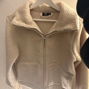 Beige teddyjacka från 157, XS - Mysig beige teddyjacka från 157 i storlek XS. Jackan har dragkedja framtill, två stora fickor och en hög krage. Perfekt för kalla dagar när du vill vara både varm och snygg. Fleece-liknande material som känns supermjukt.
