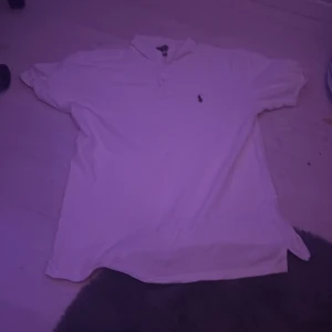 Vit pikétröja Polo Ralph Lauren L - Vit pikétröja från Polo Ralph Lauren i storlek L. Slim fit-modell med klassisk krage, knäppning framtill och broderad logga på bröstet. Tillverkad i stretchig bomullsmix som är skön och luftig. Perfekt för en clean och stilren look.