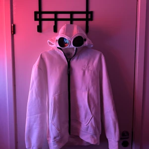 Vit hoodie med goggles från C.P. Company - Vit hoodie från C.P. Company med ikoniska goggles i huvan och broderad logga på bröstet. Tröjan har dragkedja framtill, två stora fickor och en clean, modern look. Perfekt för dig som vill sticka ut med streetwear-vibe. 
