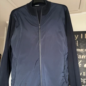 Mörkblå Cardigan John Henric S - Snygg mörkblå Cardigan från John Henric i storlek S. Jackan har en stilren design med ribbad krage och ärmar, hel dragkedja framtill och slät yta. Materialet är en mix av polyester och stickad tyg på ärmarna för extra komfort.