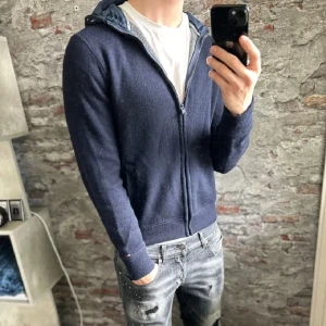 Marinblå hoodie från Tommy Hilfiger - Snygg marinblå hoodie från Tommy Hilfiger med dragkedja och huva i glansigt material. Tröjan har långa ärmar, två fickor fram och en liten logga på ärmslutet. Perfekt för dig som gillar en stilren och sportig look.