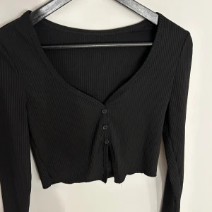 Svart ribbad croppad topp med knappar - Svart långärmad croppad topp med ribbad struktur och v-ringning. Toppen har små svarta knappar framtill.