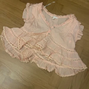 Ljusrosa volangblus med spetsdetaljer - Supersöt ljusrosa blus med volanger och spetsdetaljer. Blusen har v-ringning, korta volangärmar och knappar framtill. Perfekt för dig som gillar romantisk och bohemisk stil.