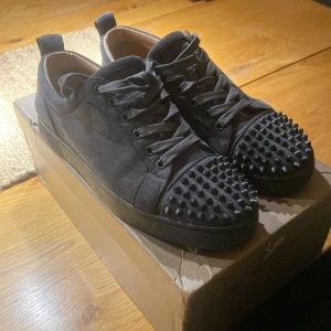 Christian Louboutin svarta sneakers med nitar - Svarta sneakers från Christian Louboutin i mocka med ikonisk röd sula och coola svarta nitar på tån. Skorna har snörning och en platt sula, perfekt för dig som vill sticka ut med en edgy vibe. Märkets logga syns på sulan.