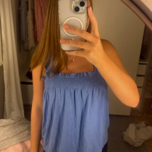 Blå linnetopp från Kappahl - Supersöt blå linnetopp från Kappahl med smock upptill och volangkant vid axelbanden. Toppen är ärmlös och har en luftig, loose passform som är perfekt för varma dagar. Tillverkad i mjuk bomull som känns skön mot huden.